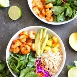 Two bowls with shrimp, avocado, brown rice, spinach, shredded carrots, purple cabbage, and diced mango, garnished with lime and fresh cilantro on a dark surface.