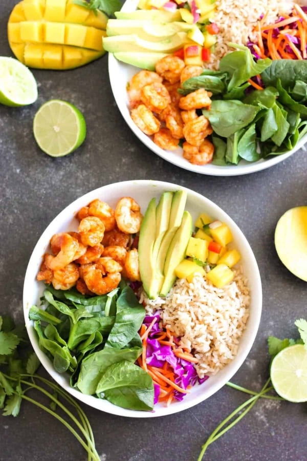 Two bowls with shrimp, avocado, brown rice, spinach, shredded carrots, purple cabbage, and diced mango, garnished with lime and fresh cilantro on a dark surface.
