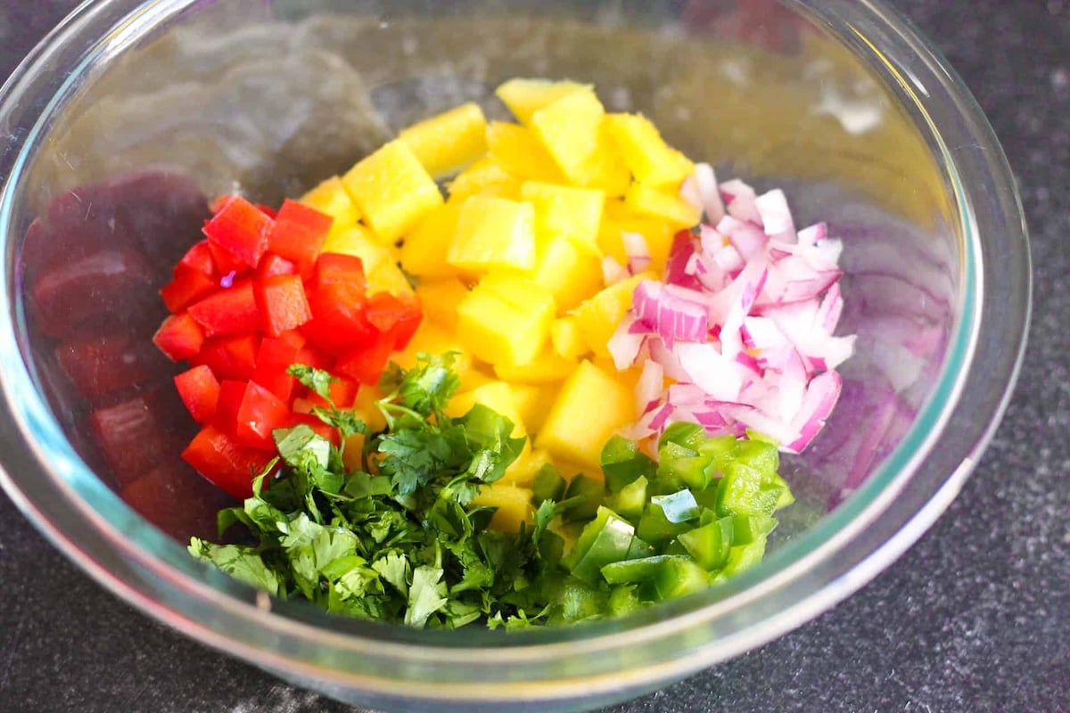 A glass bowl containing chopped red bell pepper, pineapple, red onion, green bell pepper, and cilantro on a dark countertop.