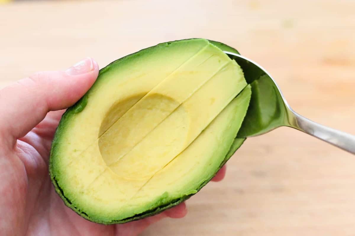 A hand holds a halved avocado with scored flesh as a spoon scoops out a section. The background is blurred.