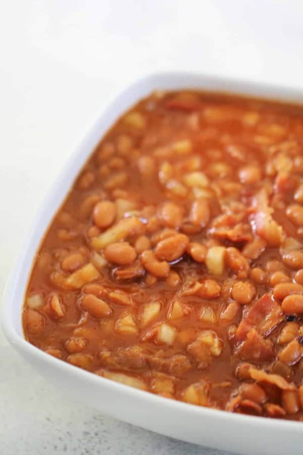 A square white dish filled with baked beans in a rich, reddish-brown sauce with visible chunks of onion and bacon.