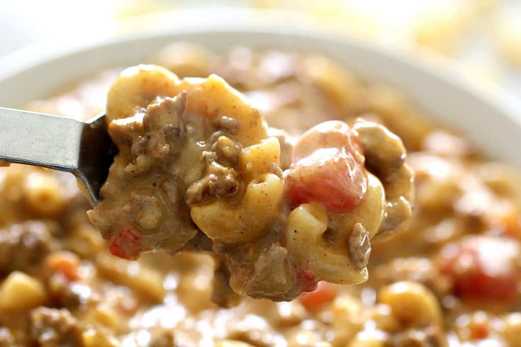 A spoon lifts a portion of creamy macaroni and ground beef with diced tomatoes from a bowl of the same dish.