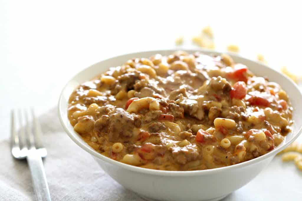 A bowl of macaroni and cheese with ground beef and diced red bell peppers, placed next to a fork on a white surface.