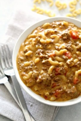 A bowl of creamy macaroni pasta mixed with ground beef and diced tomatoes, placed next to two forks on a white napkin.