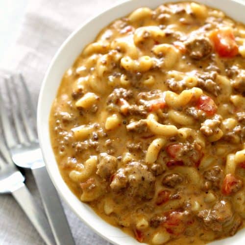 A bowl of creamy macaroni pasta mixed with ground beef and diced tomatoes, placed next to two forks on a white napkin.