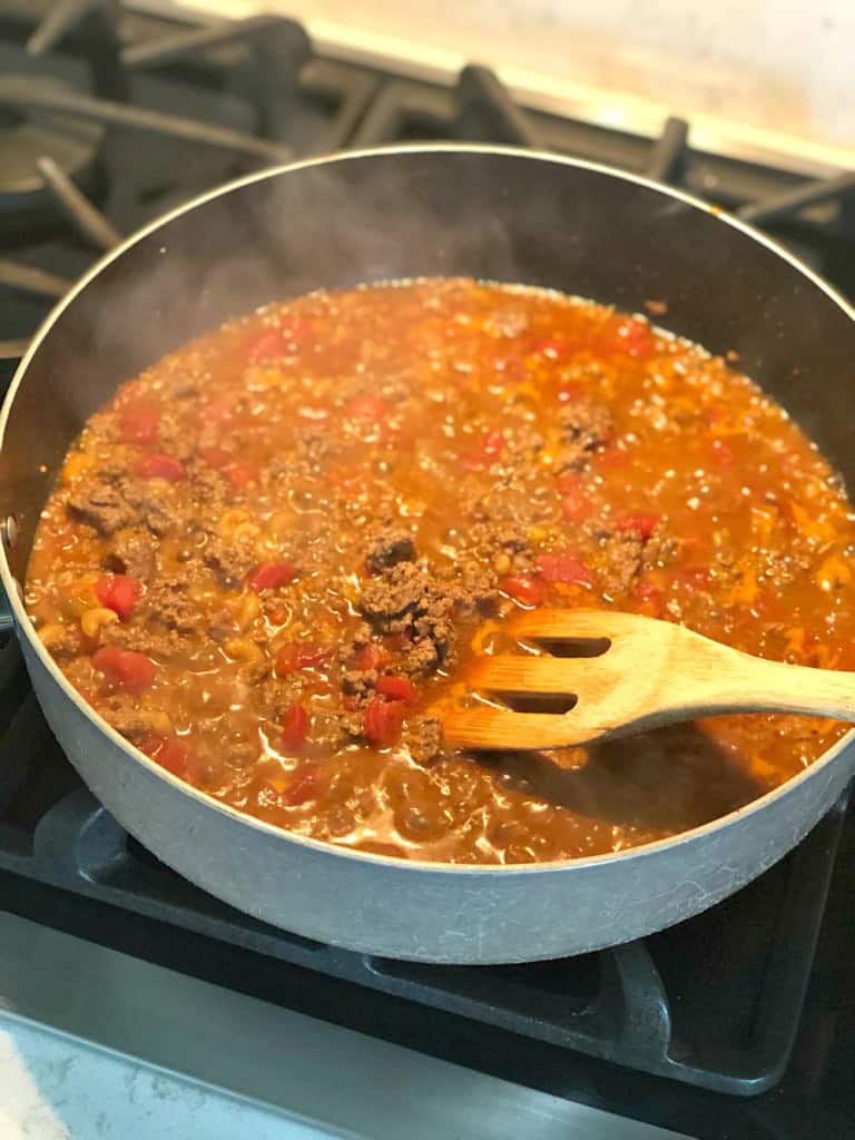 A pot of seasoned ground meat cooking with red bell peppers and tomatoes, being stirred with a wooden spoon on a stove.