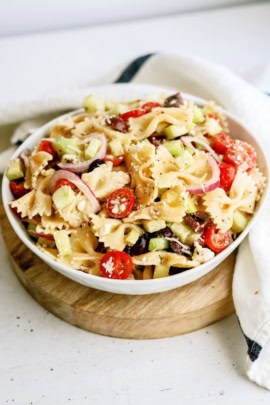A bowl of Greek Pasta Salad in a white bowl on top of a wooden saucer.