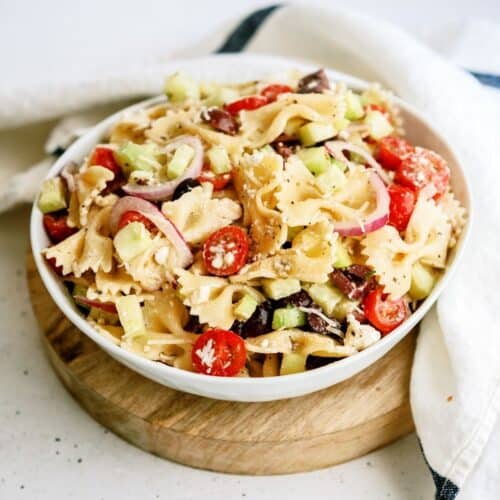 A bowl of Greek Pasta Salad in a white bowl on top of a wooden saucer.
