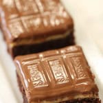 A close-up of chocolate brownie squares topped with a layer of chocolate, arranged in a row on a white plate.