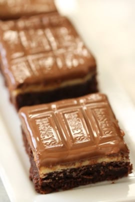 A close-up of chocolate brownie squares topped with a layer of chocolate, arranged in a row on a white plate.