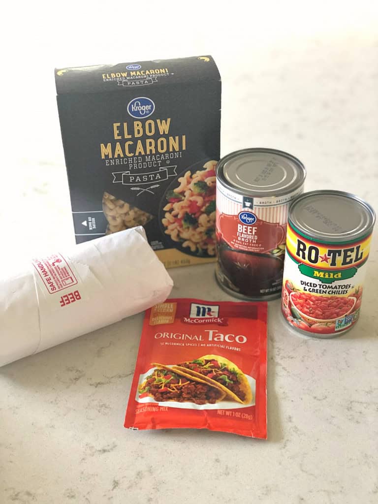 Ingredients on a counter: a box of elbow macaroni, a can of beef broth, a can of diced tomatoes and green chilies, a packet of taco seasoning, and a roll of wrapped beef.