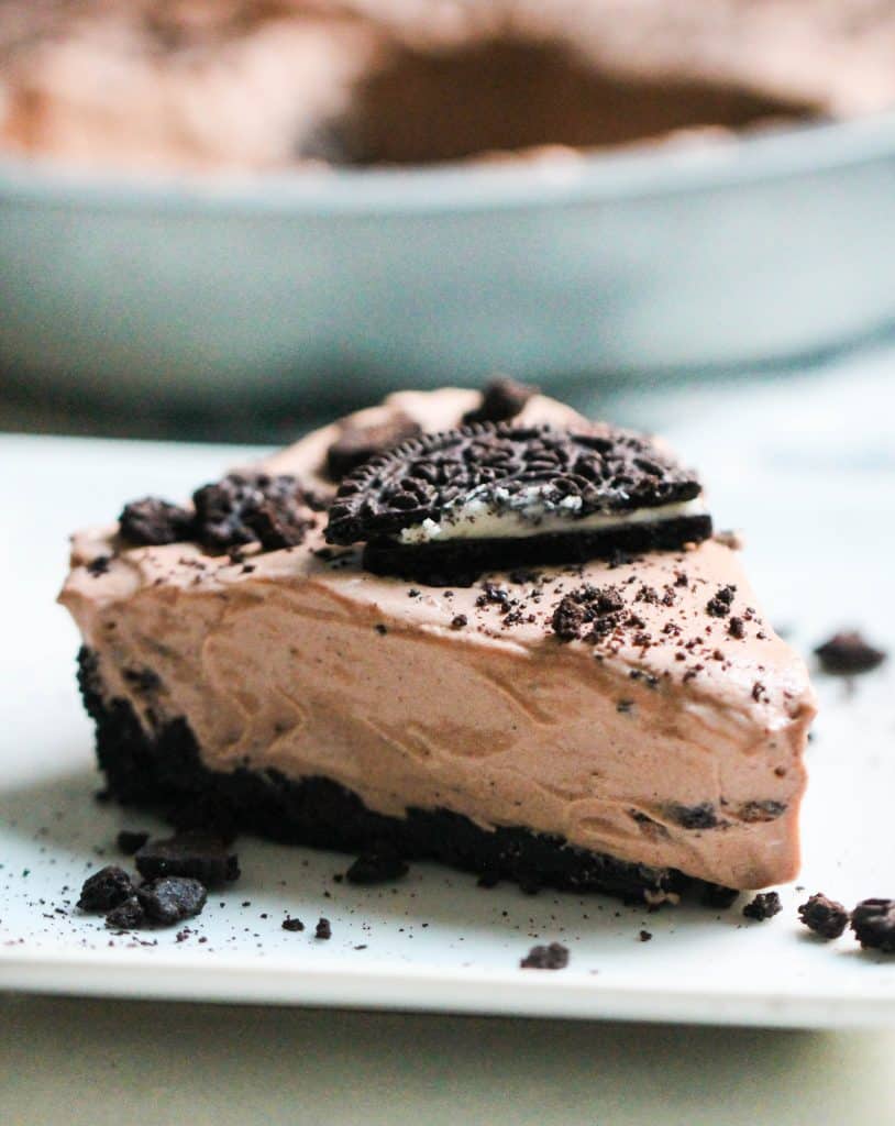 A close-up of a chocolate oreo pie topped with crumbled bits in a round metal pan on a light background with a slice missing