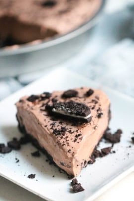A slice of chocolate cream pie topped with crushed cookies and a large cookie piece on a square white plate, with the whole pie visible in the background.