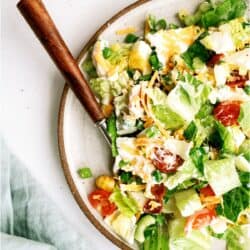 A plate of chopped salad with lettuce, cherry tomatoes, shredded cheese, hard-boiled eggs, and green onions, served with a fork on a white surface.