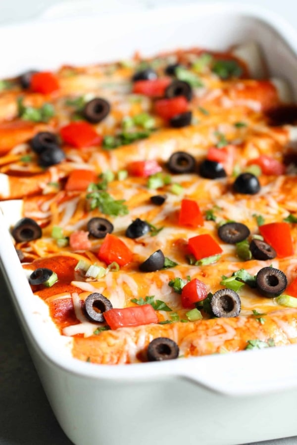 A close-up of a casserole dish filled with enchiladas, topped with melted cheese, sliced black olives, diced red bell peppers, and chopped green onions.
