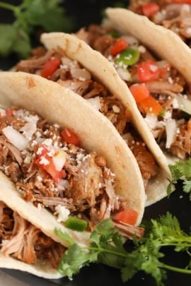 Three soft shell tacos filled with shredded meat, diced tomatoes, onions, green peppers, and topped with crumbled cheese, garnished with fresh cilantro.