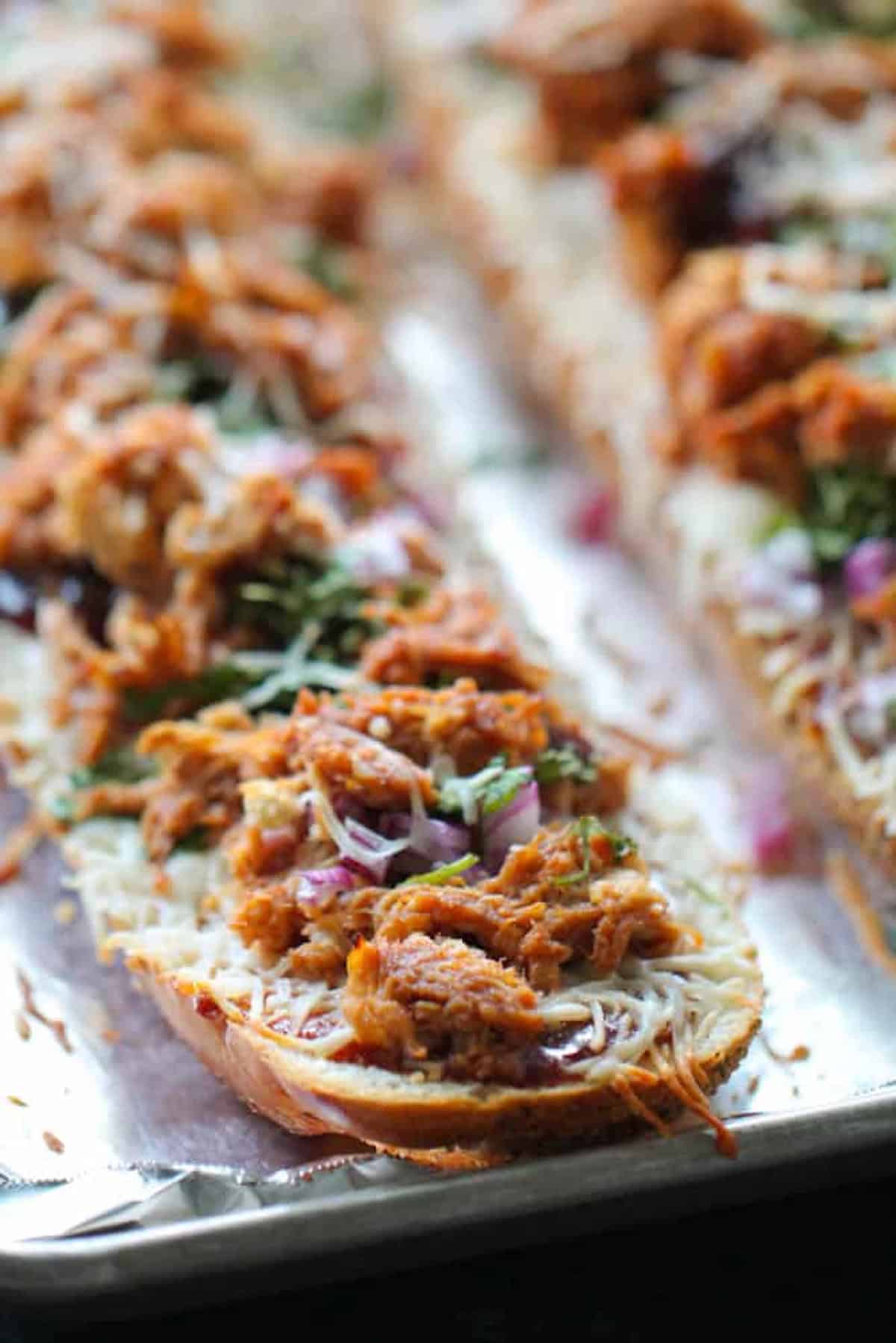 Close-up of toasted bread slices topped with shredded barbecue chicken, melted cheese, chopped red onions, and fresh herbs on a baking sheet.