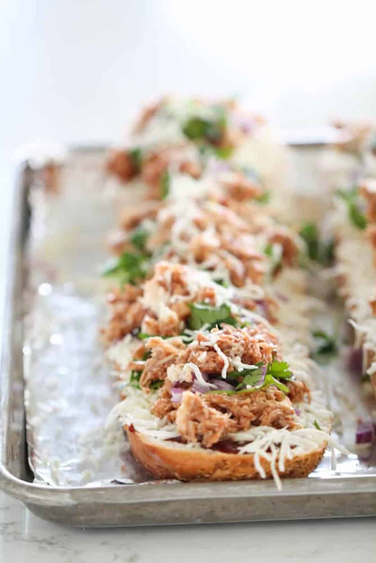 Several pieces of bread topped with shredded meat, cheese, and fresh herbs are arranged on a foil-lined baking tray.