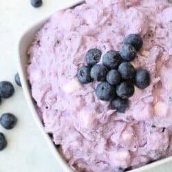 A white bowl filled with a creamy, purple blueberry salad topped with fresh blueberries, surrounded by a few scattered blueberries on a light surface.
