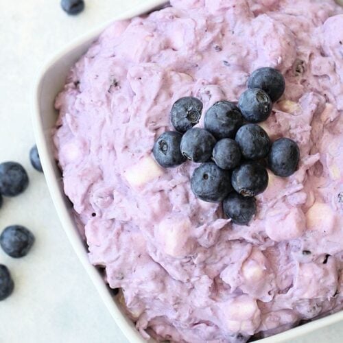 A white bowl filled with a creamy, purple blueberry salad topped with fresh blueberries, surrounded by a few scattered blueberries on a light surface.