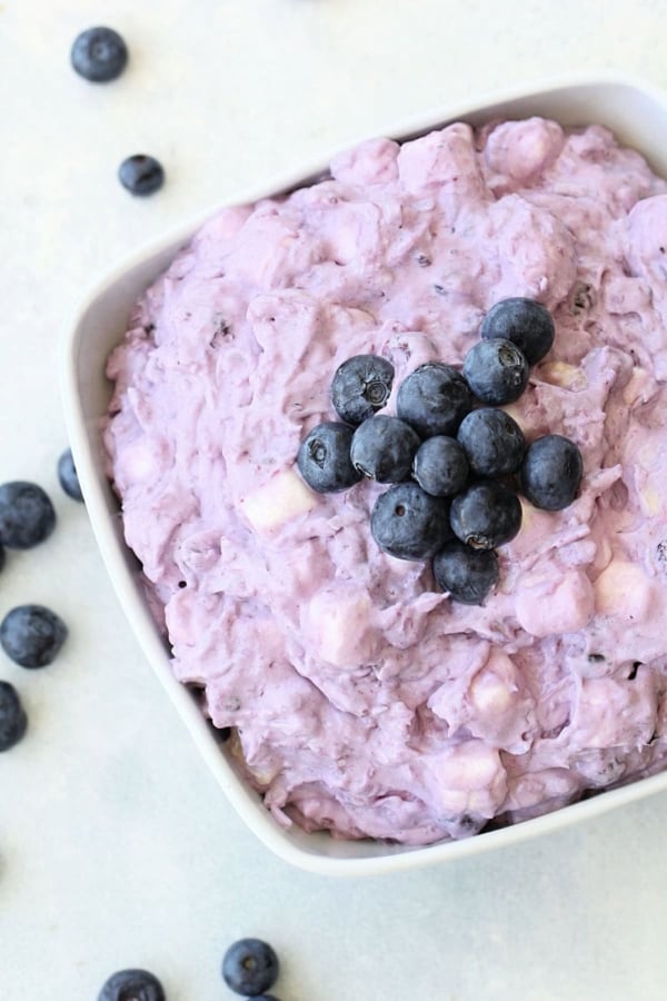A white bowl filled with a creamy, purple blueberry salad topped with fresh blueberries, surrounded by a few scattered blueberries on a light surface.
