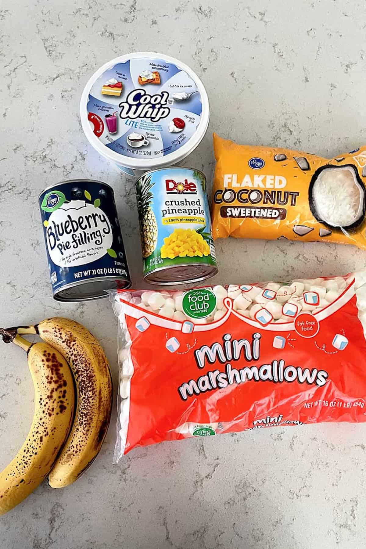 Assorted dessert ingredients on a counter, including Cool Whip, blueberry pie filling, crushed pineapple, flaked coconut, mini marshmallows, and two ripe bananas.
