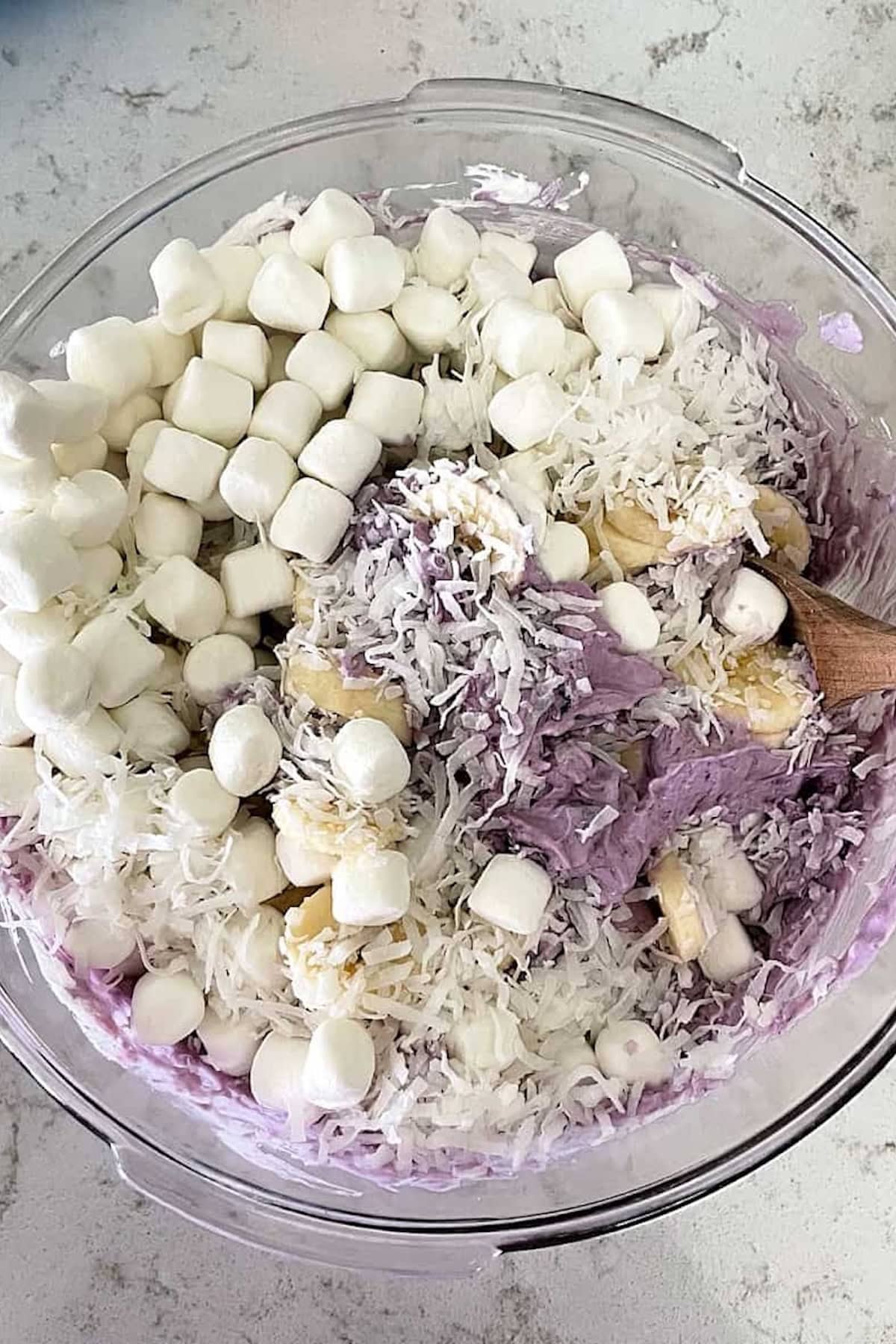 A glass bowl containing whipped purple mixture, mini marshmallows, shredded coconut, chopped nuts, and a wooden spoon.