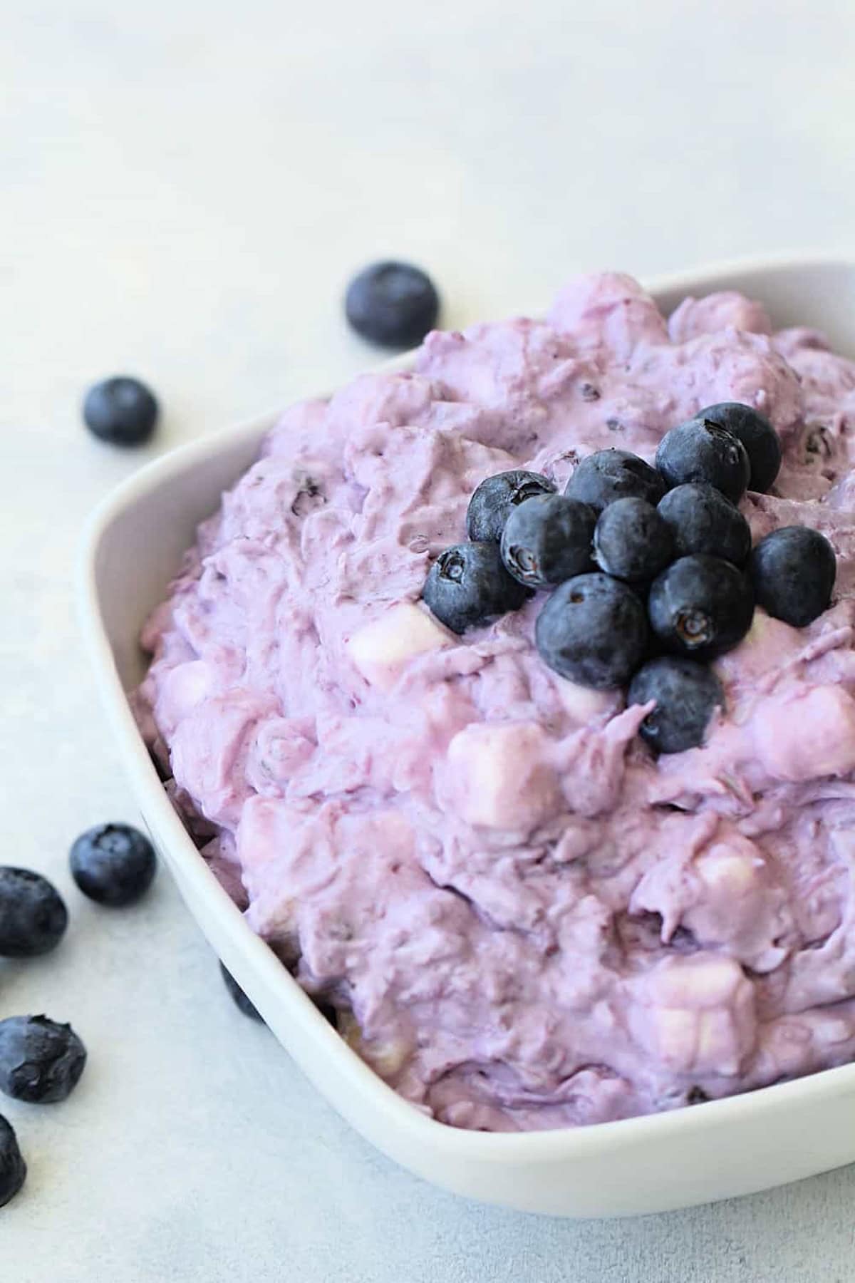 A bowl of purple fruit salad with mini marshmallows, topped with fresh blueberries, with a few loose blueberries on the table.