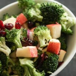 A bowl of broccoli salad with chopped apples, dried cranberries, and red onion pieces on a dark surface.