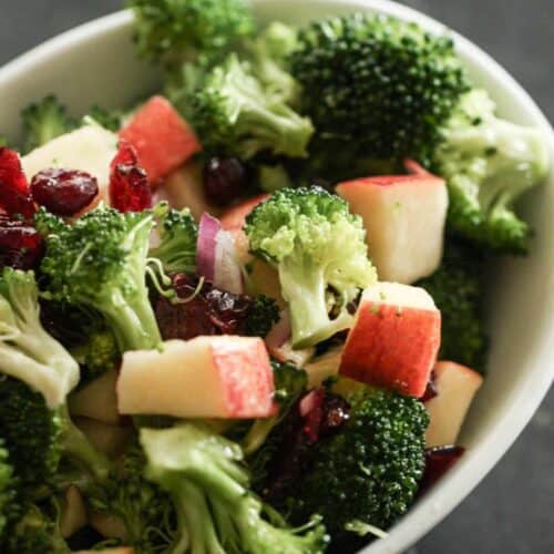 A bowl of broccoli salad with chopped apples, dried cranberries, and red onion pieces on a dark surface.
