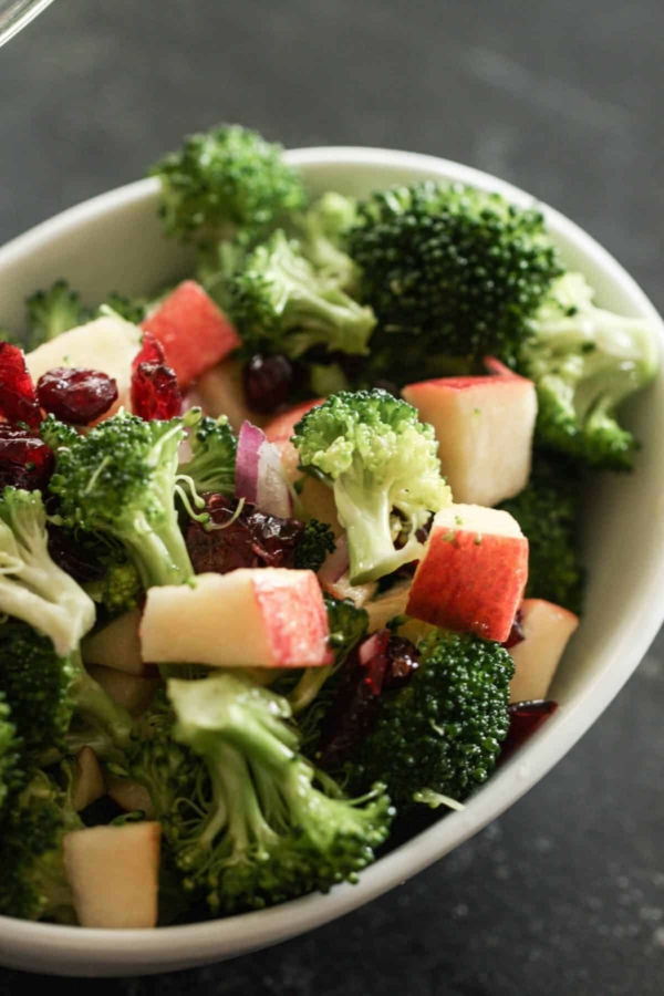A bowl of broccoli salad with chopped apples, dried cranberries, and red onion pieces on a dark surface.