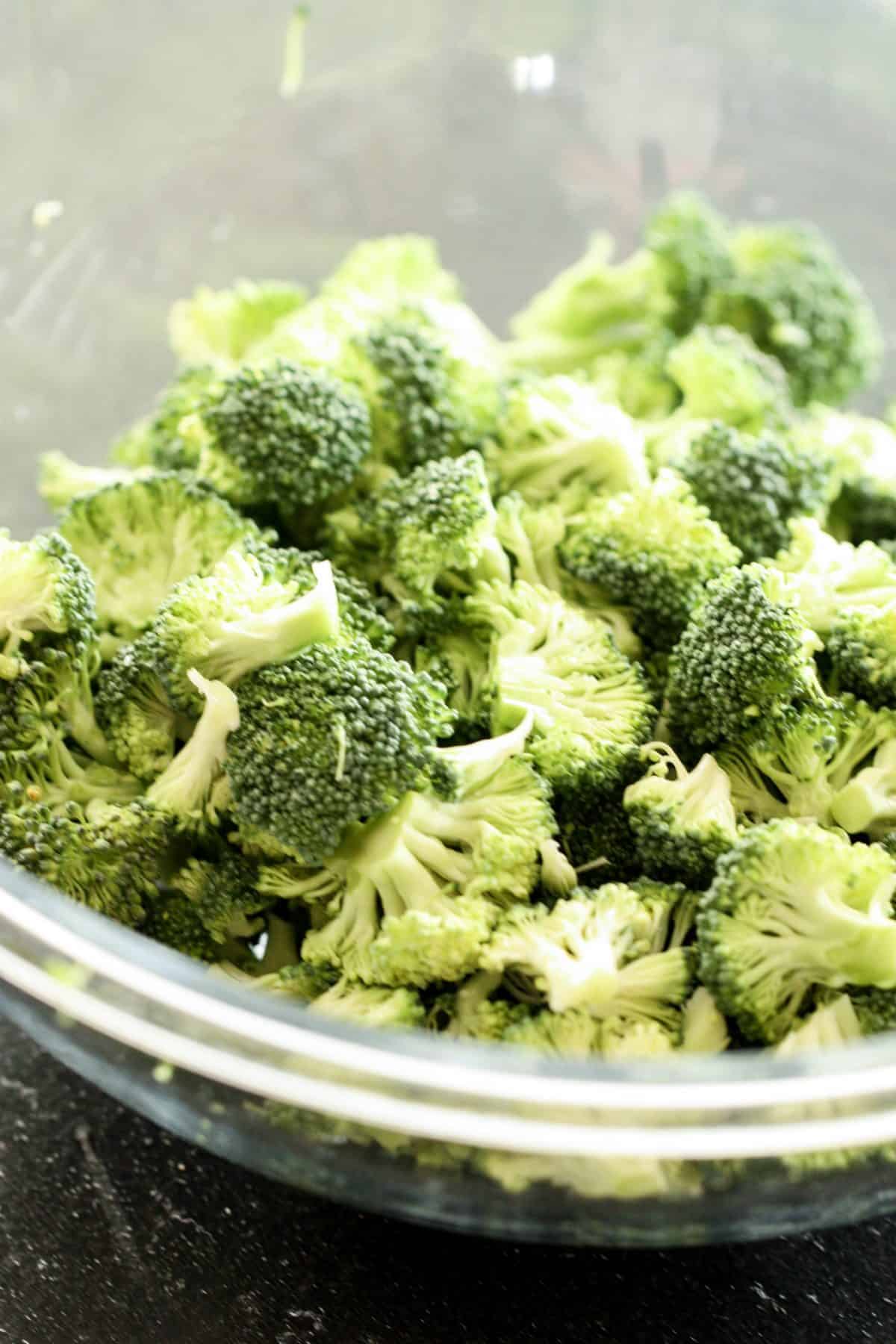 A clear glass bowl filled with fresh, uncooked broccoli florets on a dark surface.