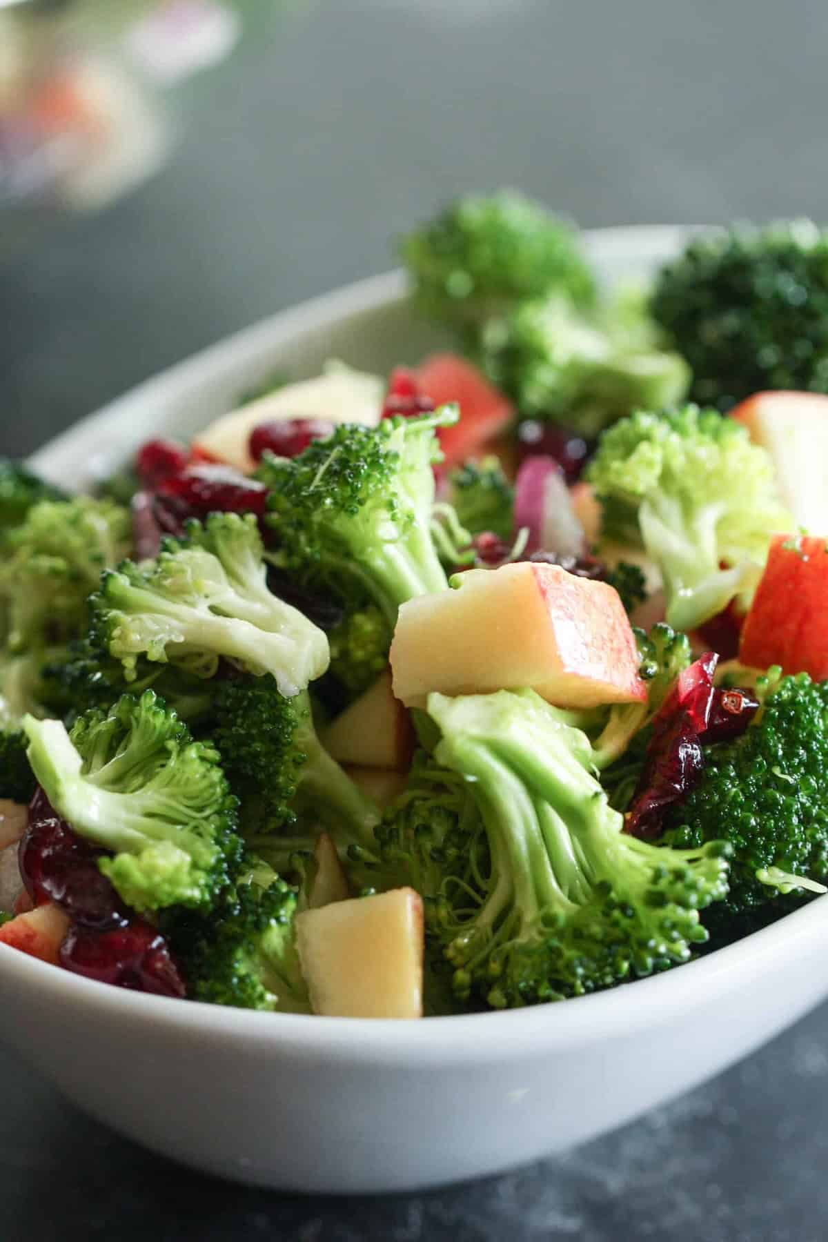 A white bowl filled with a fresh broccoli salad, featuring chopped broccoli, apple pieces, red onion, and dried cranberries.
