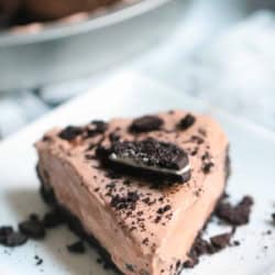 A slice of chocolate cream pie topped with crushed cookies and a large cookie piece on a square white plate, with the whole pie visible in the background.