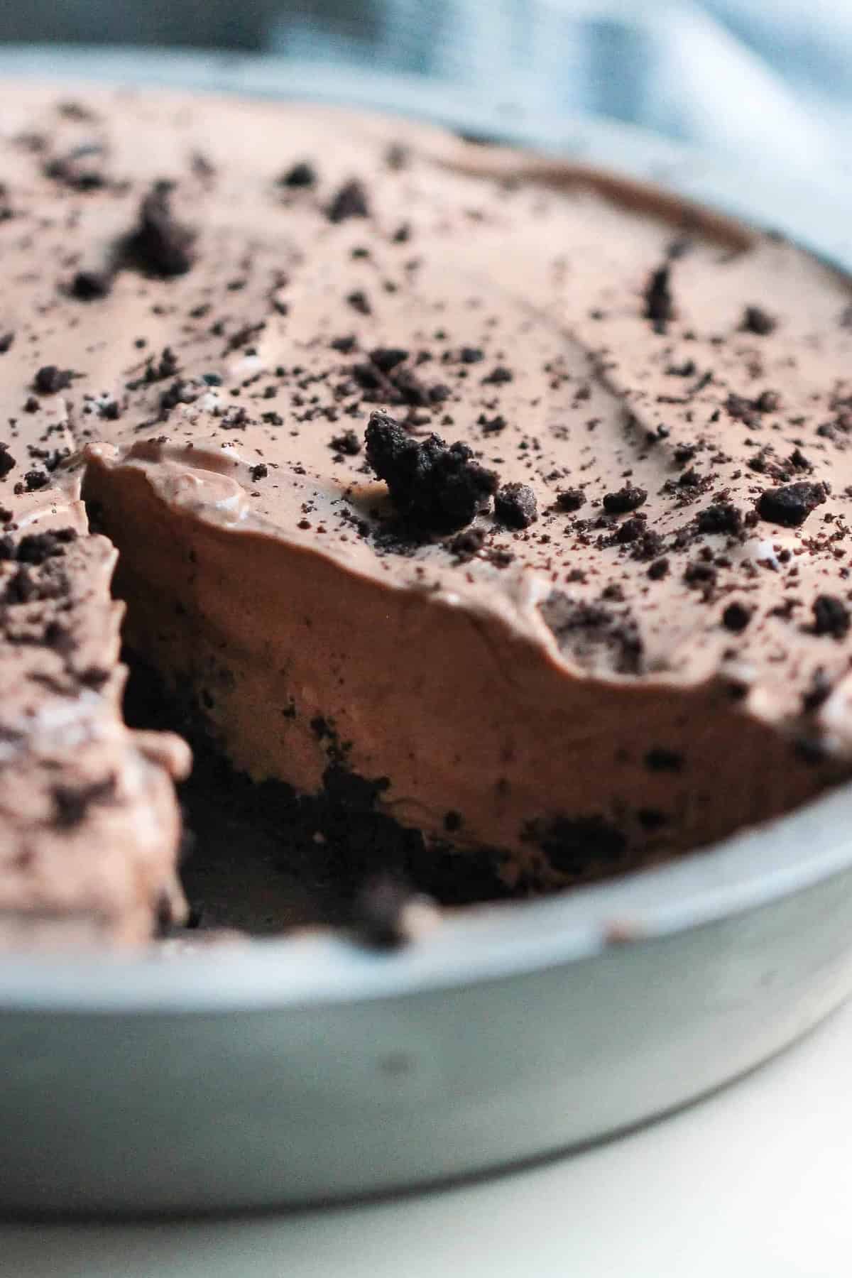 A close-up of a chocolate oreo pie topped with crumbled bits in a round metal pan on a light background with a slice missing