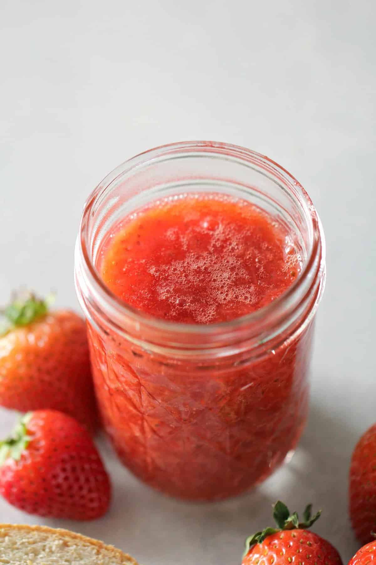 A glass jar filled with strawberry jam sits on a light surface, surrounded by fresh strawberries.