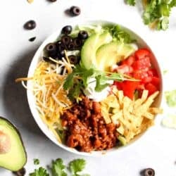 A bowl of taco salad with ground meat, shredded cheese, sliced olives, avocado, tomato, lettuce, tortilla strips, and a dollop of sour cream, surrounded by fresh ingredients.