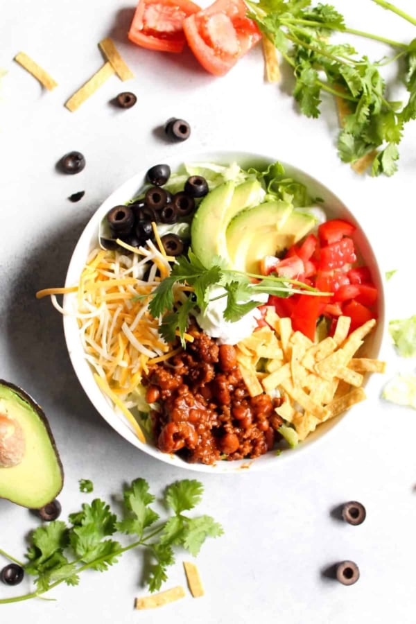 A bowl of taco salad with ground meat, shredded cheese, sliced olives, avocado, tomato, lettuce, tortilla strips, and a dollop of sour cream, surrounded by fresh ingredients.