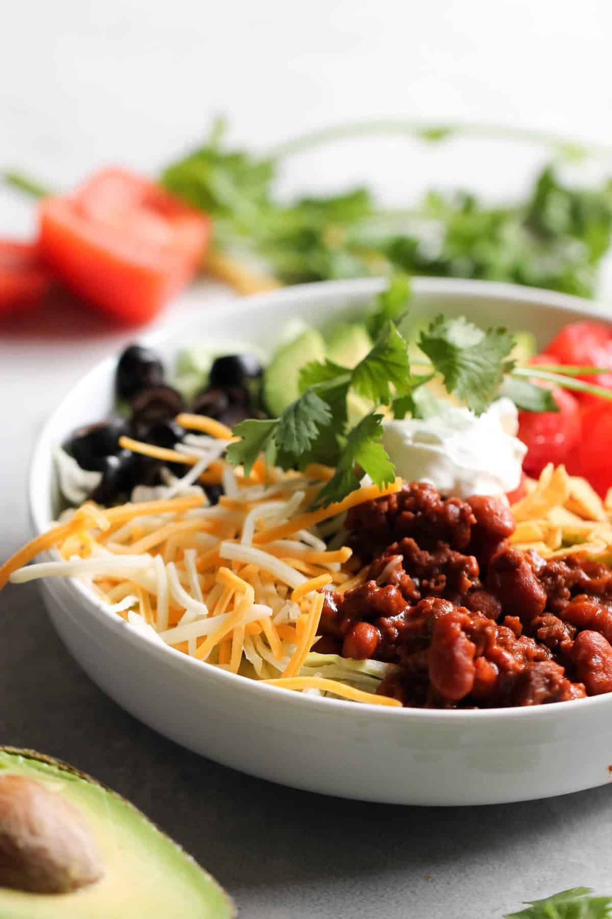 A bowl containing ground meat with beans, shredded cheese, black olives, sour cream, cilantro, sliced cucumber, and tomato, with avocado and more vegetables in the background.