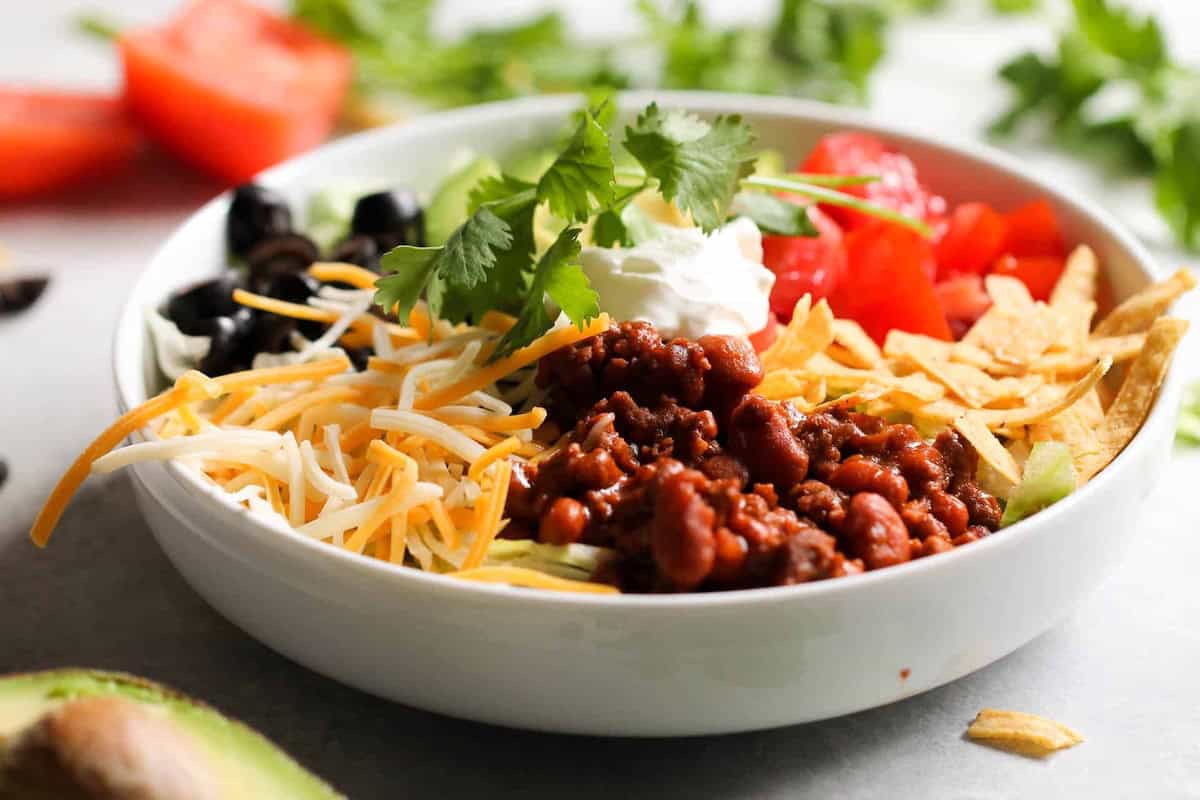 A bowl with chili beans, shredded cheese, black olives, tomato, tortilla strips, avocado, sour cream, and cilantro, arranged in sections on a white plate.