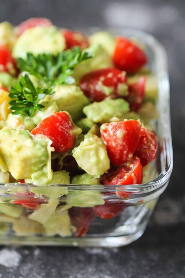 A close-up of a fresh salad in a glass container, featuring diced avocado, cherry tomatoes, and garnished with parsley.