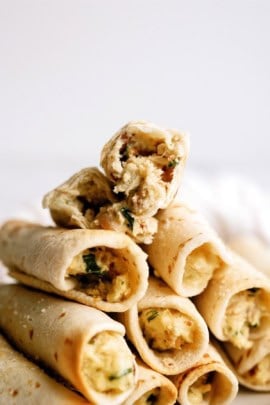 A stack of rolled tortillas filled with a mixture of scrambled eggs, herbs, and likely meat, with one tortilla on top broken in half to display the filling.