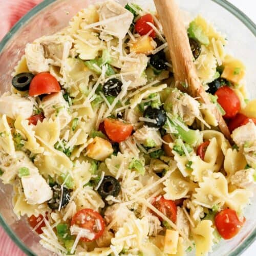 A bowl of pasta salad with bowtie pasta, cherry tomatoes, chicken, black olives, cheese, and greens, mixed with a wooden spoon.