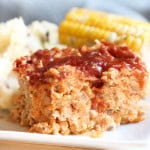 Close-up of a plate containing a slice of meatloaf topped with sauce, mashed potatoes, and a cob of corn in the background.
