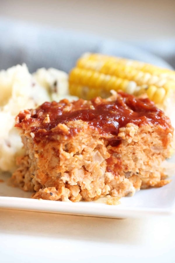 Close-up of a plate containing a slice of meatloaf topped with sauce, mashed potatoes, and a cob of corn in the background.