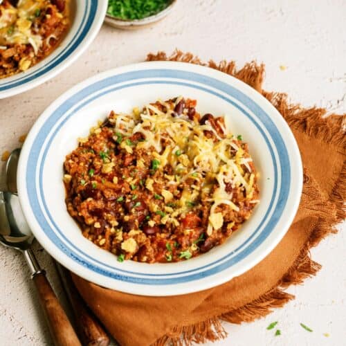 A bowl of Mom's Slow Cooker Chili topped with shredded cheese and chopped green herbs, placed on a brown napkin with a spoon beside it. Another partially visible bowl and a small dish of herbs are in the background.