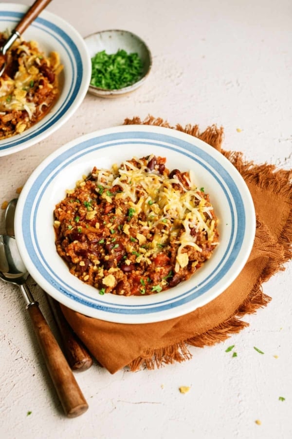 A bowl of Mom's Slow Cooker Chili topped with shredded cheese and chopped green herbs, placed on a brown napkin with a spoon beside it. Another partially visible bowl and a small dish of herbs are in the background.