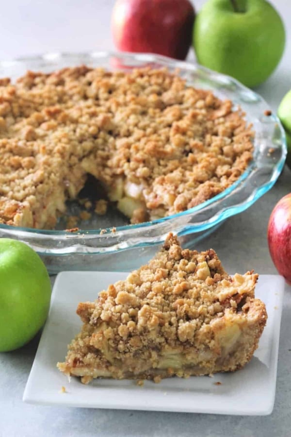 A pie with a crumbly topping is in a glass dish. A slice has been cut out and placed on a white plate in the foreground. Several whole apples are placed around the pie.