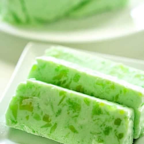 Three rectangular slices of green gelatin dessert with visible fruit pieces are arranged on a white plate, with more dessert in the background.