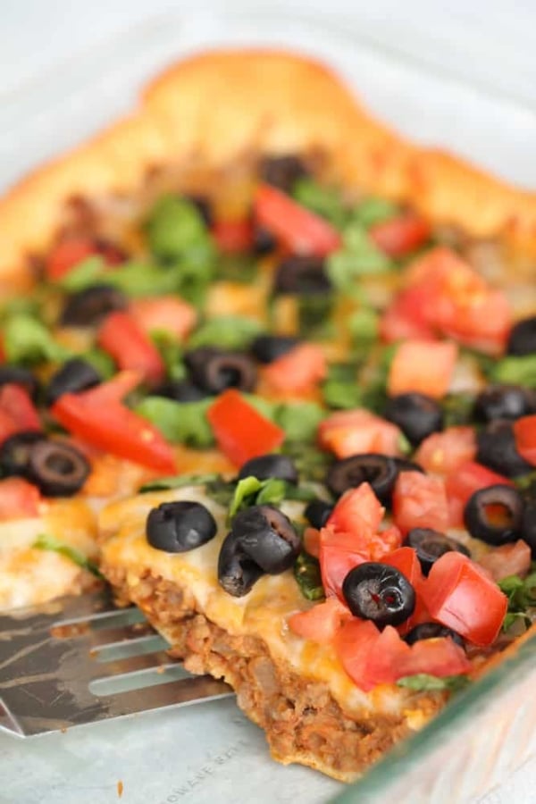 Close-up of a freshly baked pizza topped with diced tomatoes, black olives, chopped greens, and melted cheese in a glass baking dish. A metal spatula is partly visible underneath the pizza.